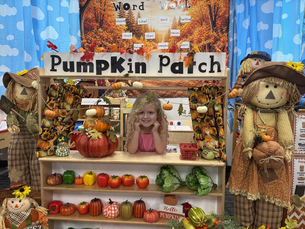 A child sits in front of a pumpkin-themed play market, surrounded by autumn decorations, pumpkins, and vegetables.