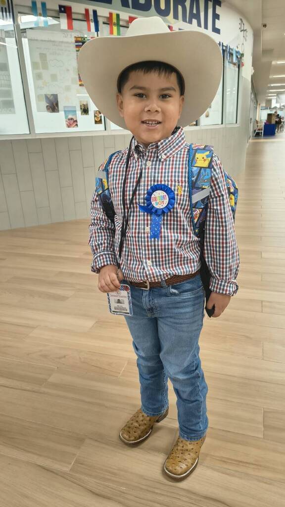 A young boy wearing a cowboy hat and plaid shirt with a backpack and blue ribbon. He stands in a hallway with wooden floors and walls.