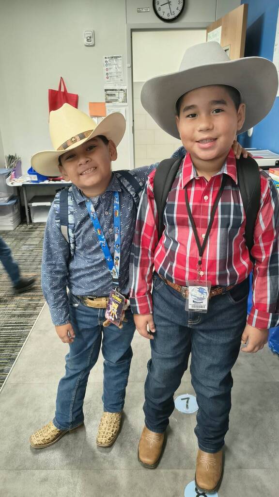 Two boys in cowboy hats and boots, one in a blue shirt, the other in a red plaid shirt, posing in a classroom.