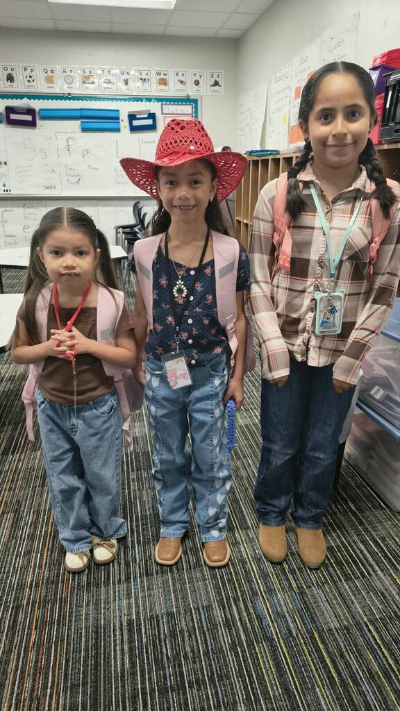 Three young girls stand together in a classroom. One wears a red cowboy hat. Behind them is a whiteboard.