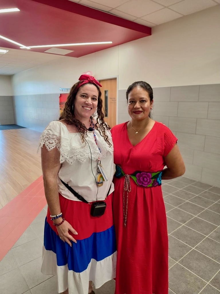 Two women stand in a hallway. One wears a white, blue, and red dress. The other wears a red dress. They are next to each other.