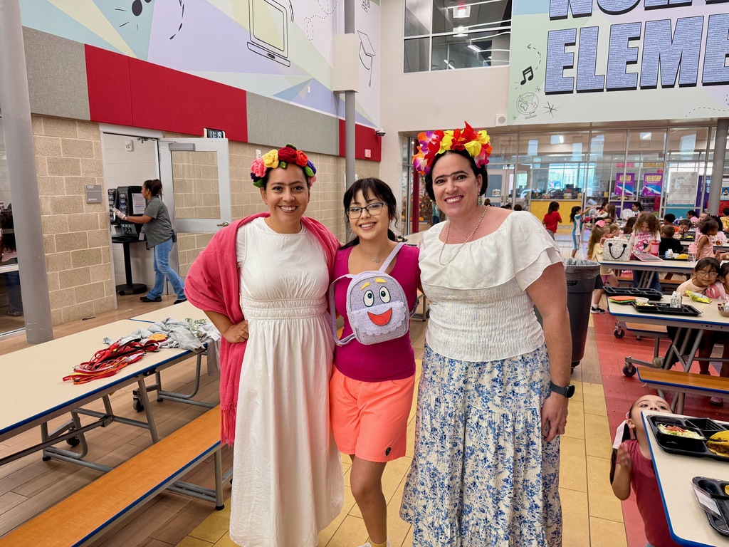 Three women stand in a school cafeteria. They wear colorful dresses and floral headbands. A white table and benches are behind them.