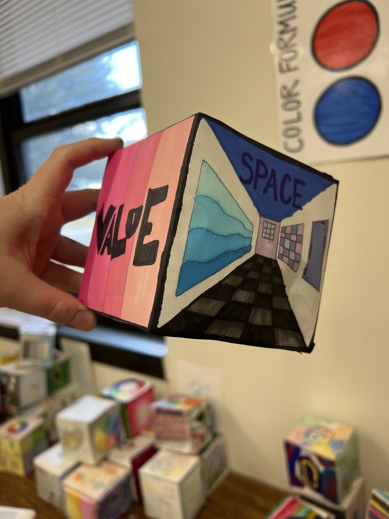 • A close-up photograph of a person's hand holding a paper cube project, showing two different faces from the previous angle. The right face is labeled "SPACE" at the top in large blue block letters, featuring a drawing of a checkered hallway using one-point perspective to create depth. The left face is colored with a pink-to-red gradient background and features the word "VALUE" written in bold, stylized black block letters. The background shows a classroom setting with blurred stacks of other paper cubes.