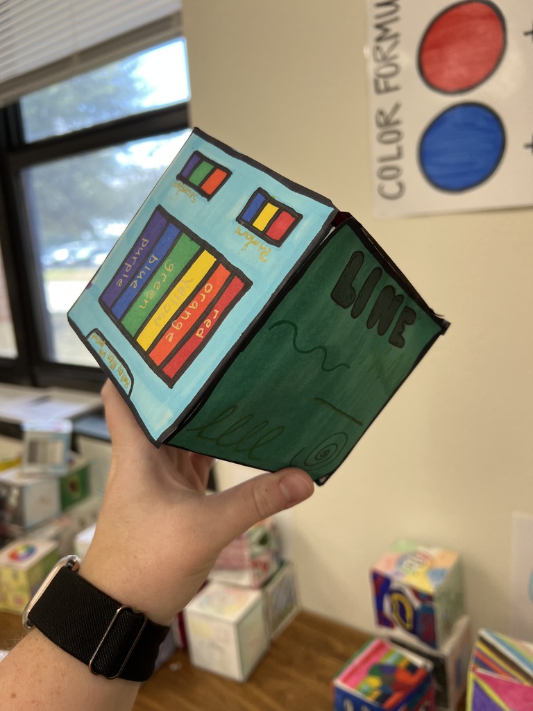 A close-up photograph of a person's hand holding a three-dimensional paper cube project in a classroom setting. The visible sides of the cube showcase different art concepts drawn with markers. One side features a blue background displaying a vertical spectrum of color bars labeled "red," "orange," "yellow," "green," "blue," and "purple," along with two smaller color-combination squares at the top. The adjacent side is colored green and displays the word "LINE" at the top in large, black block letters, with examples of different line styles drawn below it. In the background, classroom elements such as windows, desks, stacks of other student art cubes, and a "COLOR FORMULA" educational poster are visible.