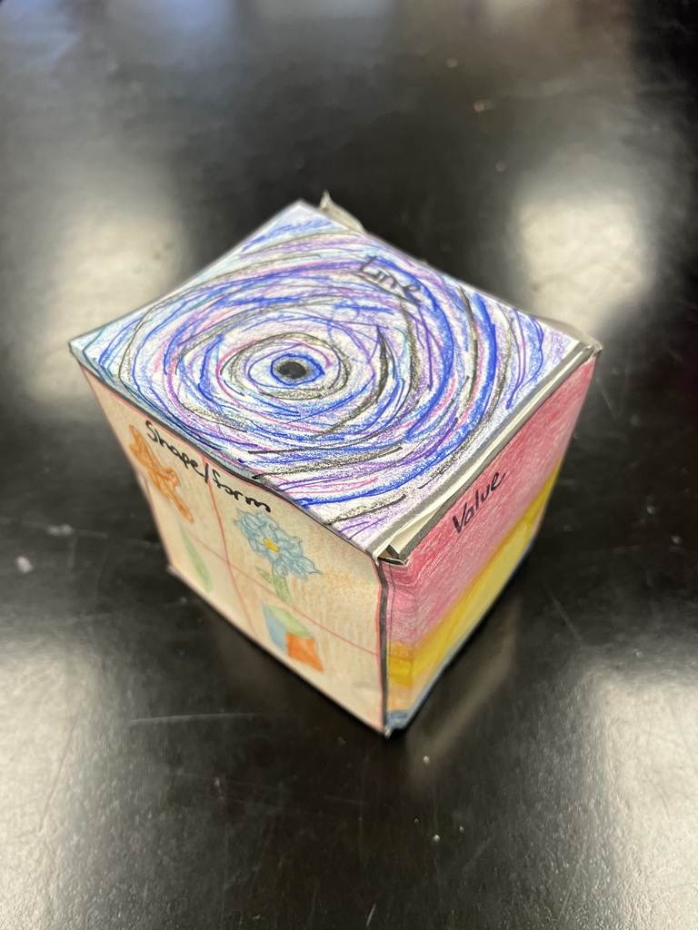 • A photograph of a different paper art cube resting on a dark black tabletop. The top face features a dense, multi-colored spiral drawing in blue, purple, and black colored pencils, labeled "Line." The left face shows drawings of flowers labeled "Shape/Form," and the right face features a pink-to-yellow gradient labeled "Value."