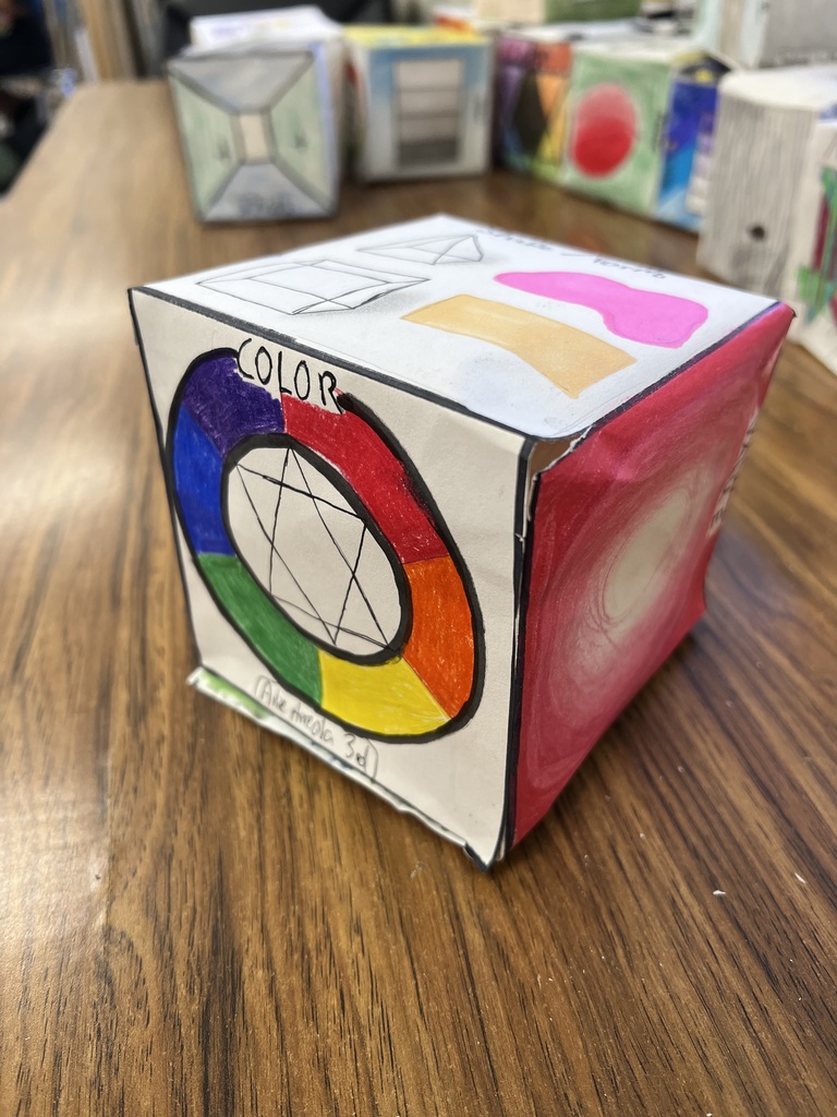 • A photograph of the same paper cube from a slightly rotated perspective on the wooden desk. The main face in view features a hand-drawn color wheel labeled "COLOR" at the top, displaying primary and secondary colors in a ring around a star geometric pattern. The red "VALUE" gradient face is visible on the right side. The top face displays geometric forms and organic shapes in soft pink and beige colored pencils.