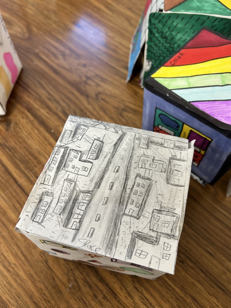 • A close-up photograph looking down at a paper art cube decorated entirely with a black-and-white graphite pencil drawing. The top face features a detailed birds-eye view of a city street with cars, buildings, and sidewalks, labeled "Space" at the bottom. Other colorful art cubes sit on the wooden desk in the background.