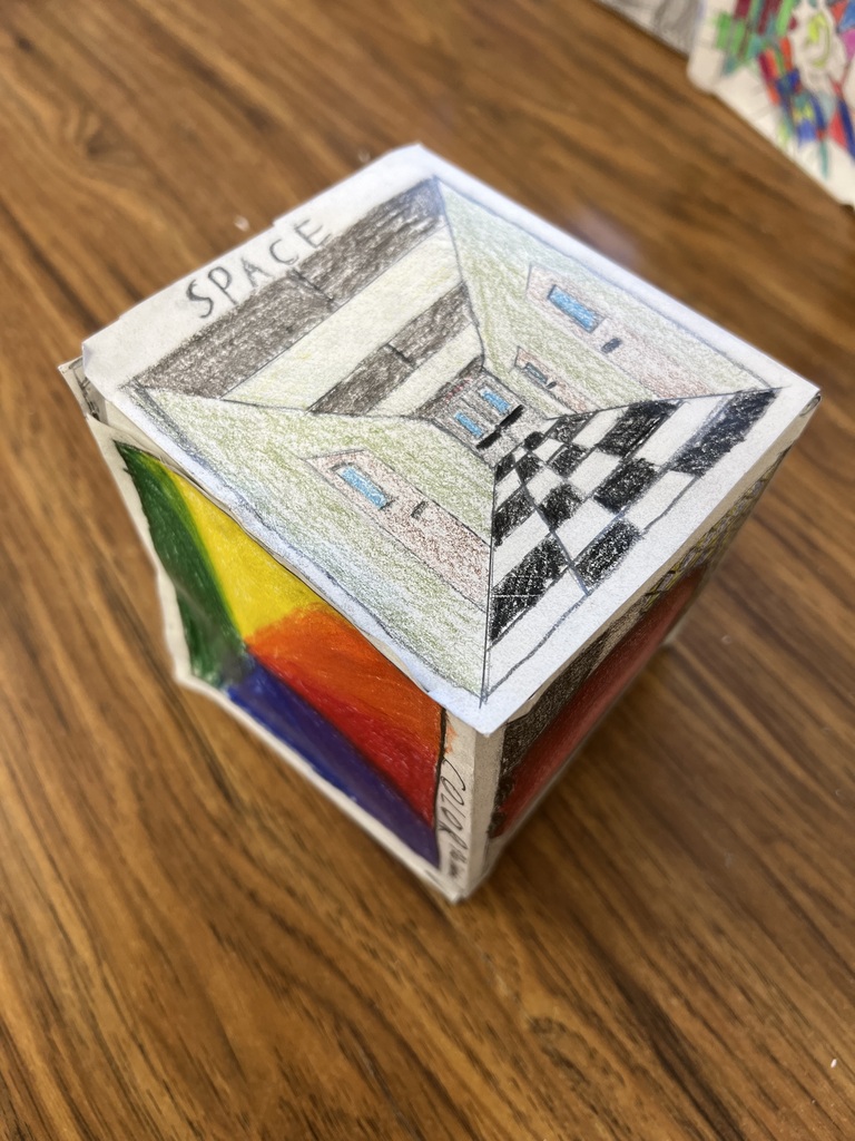 • A photograph of the same cube from Image 5, rotated to show different sides. The top face features a one-point perspective drawing of a checkered floor hallway under the label "SPACE." The left face shows a bold, multi-colored section with triangular blocks of color. The right face, labeled "COLOR," is partially visible.