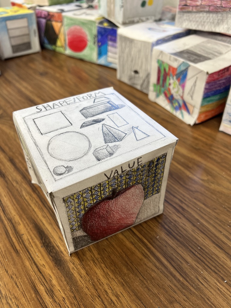 • A photograph of the cube from Image 5 and 6, rotated to showcase the "SHAPE/FORM" face on top. It contains pencil drawings of 2D shapes (square, circle) and their 3D form counterparts (cube, cylinder, pyramid, sphere). The front face features a shaded drawing of an apple against a patterned background under the label "VALUE."