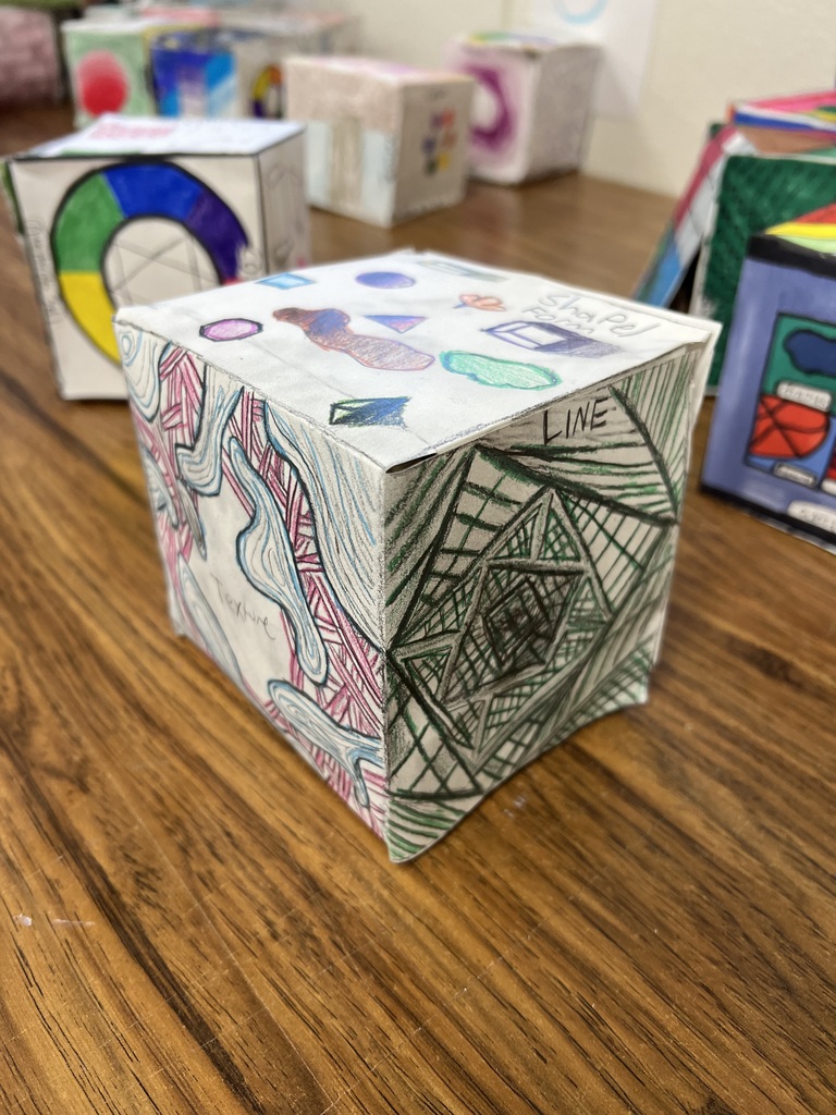 • A photograph of another paper art cube on a wooden desk. The top face contains small sketches of geometric and organic shapes labeled "Shape/Form." The front-left face features a detailed, multi-colored abstract pattern labeled "Texture." The front-right face features an intricate, green geometric line pattern labeled "LINE." Other art projects are scattered in the background.