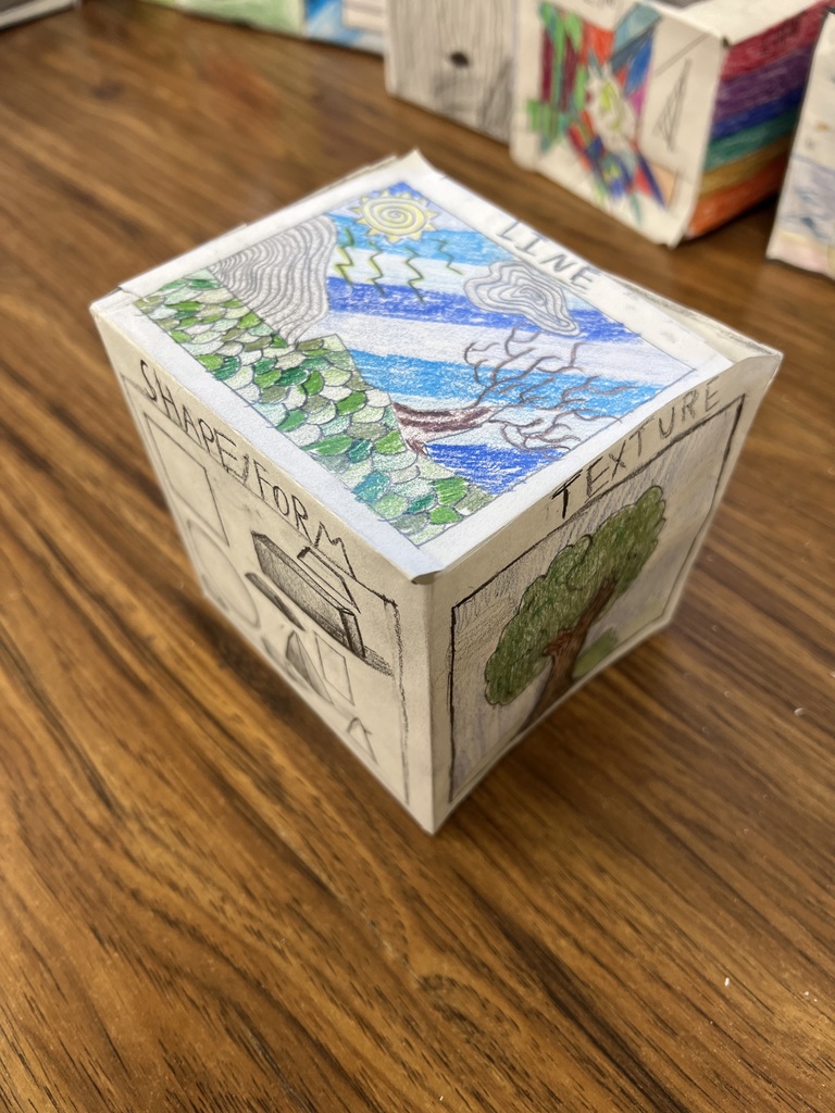 A photograph of a different paper art cube on a wooden desk. The top face features a colored pencil landscape drawing showing a sun, mountains, a tree, and water, with the word "LINE" written near the top edge. The left face shows drawn geometric figures labeled "SHAPE/FORM." The right face features a drawing of a tree, labeled "TEXTURE" along the top edge.