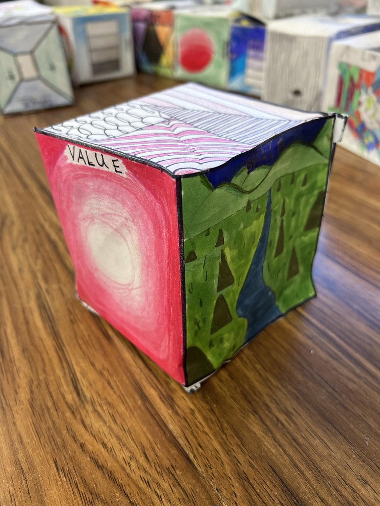 • A high-angle photograph of a paper art cube resting on a wooden desk. The front-left face features a smooth red gradient that fades to white in the center, labeled "VALUE" in small print at the top left. The front-right face features a green landscape drawing of a river winding through small triangular trees. The top face shows a pattern of wavy, pink-striped lines alongside straight, parallel blue lines. Other student art cubes are lined up in the background.