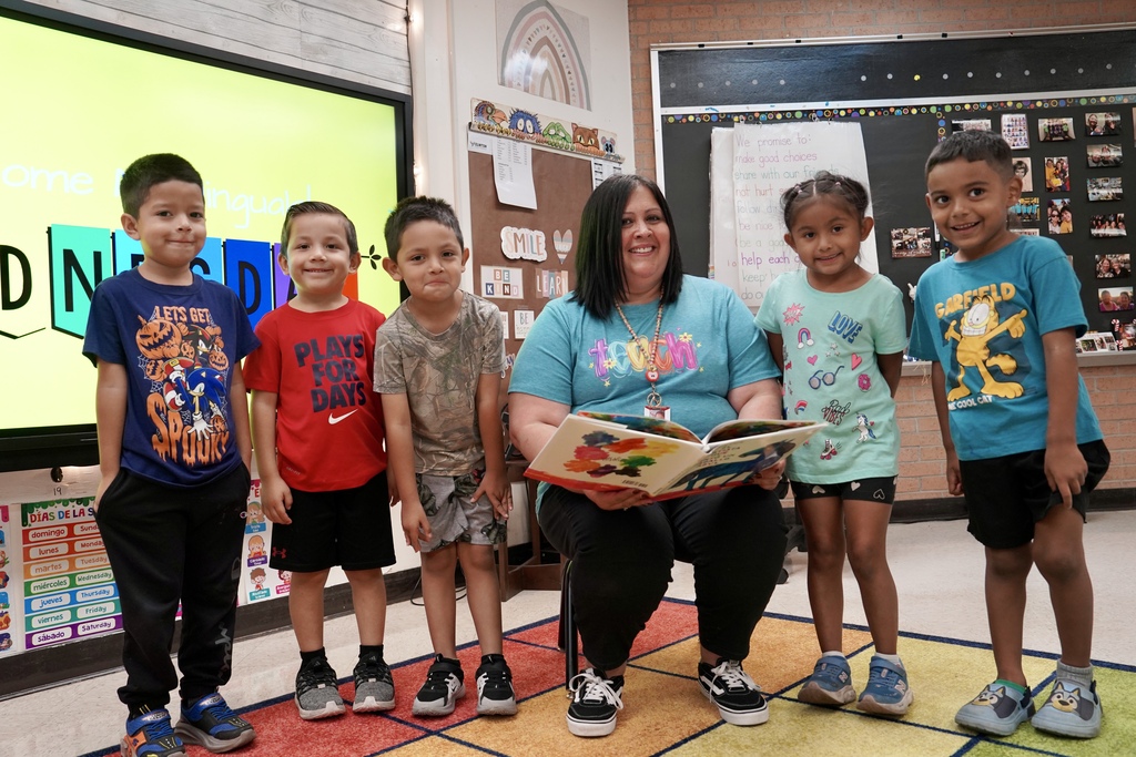 5 students standing beside their teacher reading a book in Spanish