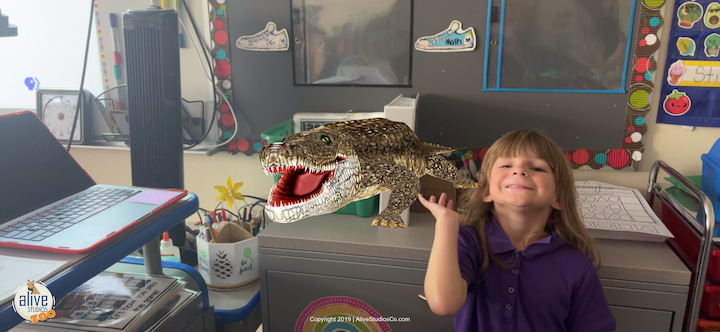 A young student smiles while holding up an image of an alligator.
