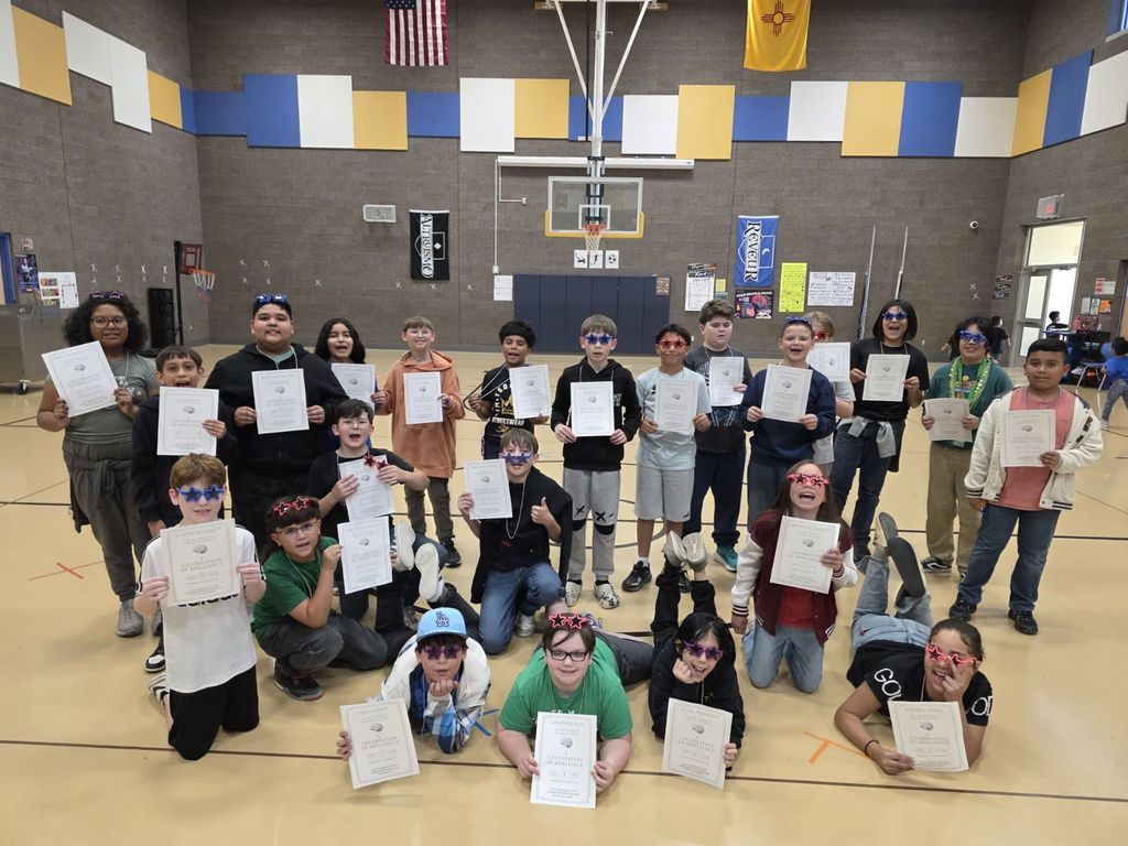 A group of people, adults and children, inside a gym, holding certificates, smiling, and posing for a photo.