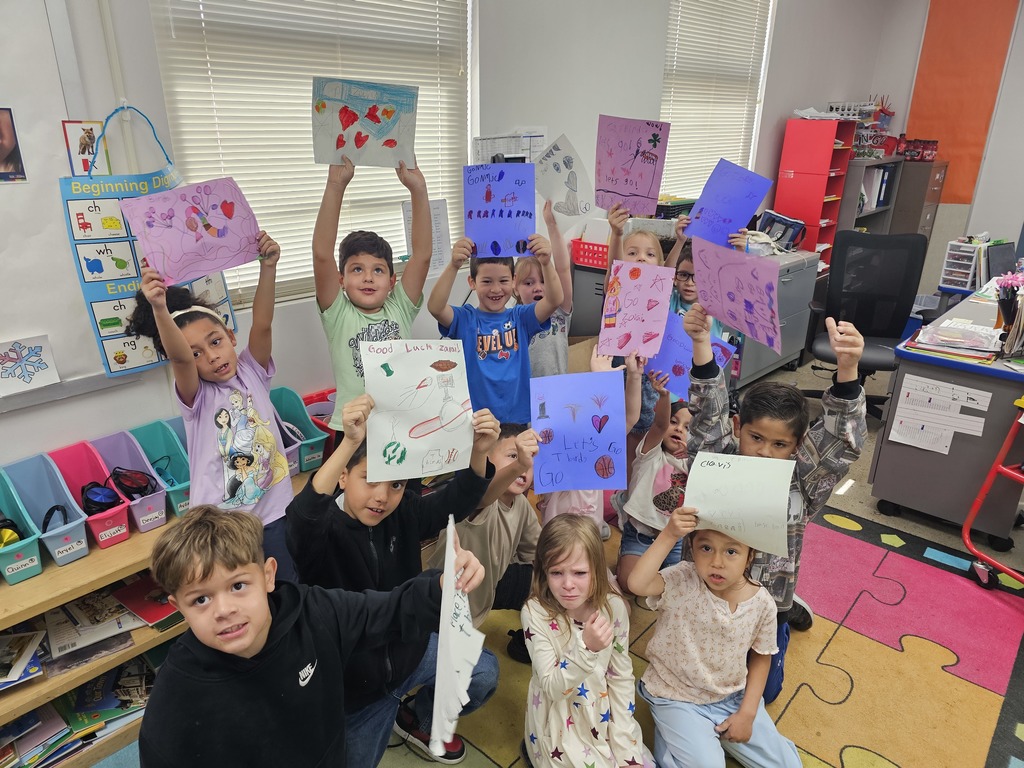 Several children inside a classroom hold up heart-shaped artwork. Behind them, shelves and storage boxes are visible.
