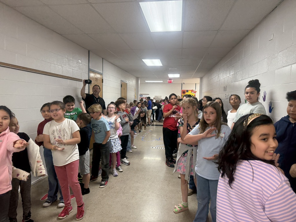 A group of children in school attire line up in a hallway with white walls and ceiling lights.