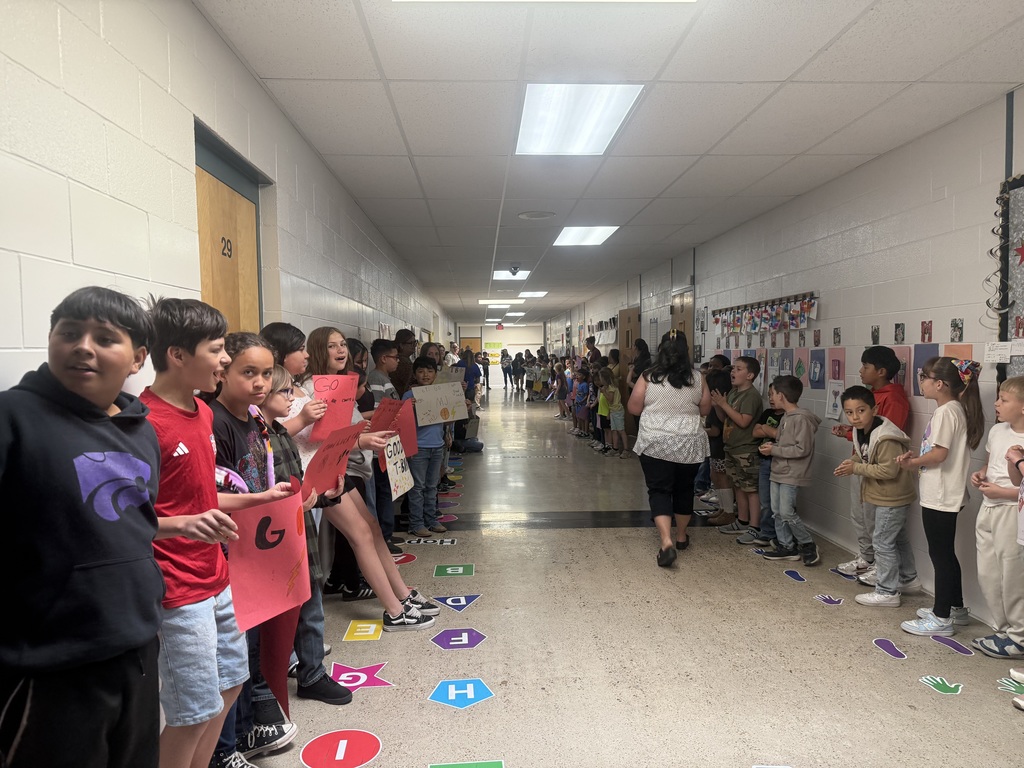 Children stand in line, holding signs. They face away from a hallway with white walls and ceiling lights.