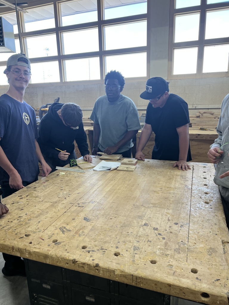 Five individuals are gathered around a large wooden table, possibly working on a project. They are in a room with large windows.
