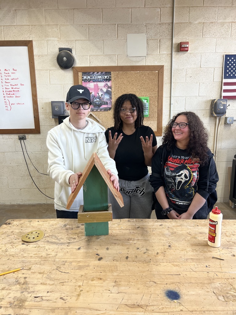 Three individuals in a workshop, one holding a wooden birdhouse, and two others gesturing peace signs. A white wall with various objects and a flag is behind them.