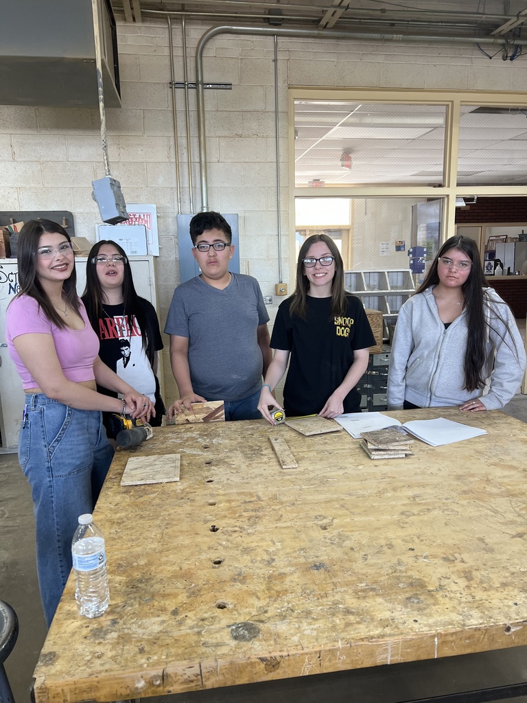 Five individuals stand around a wooden table, two holding tools and three holding papers. Background shows a room with a large window and a metal pipe.