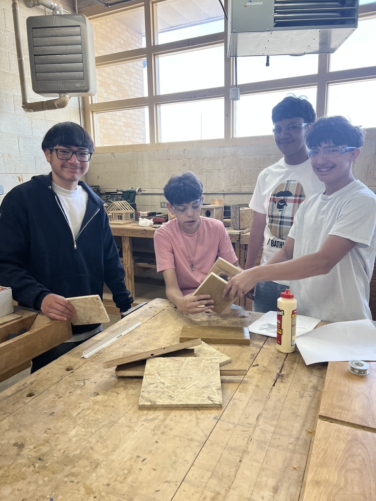 Four people in a workshop, working on a wooden project at a table, with wood pieces, tools, and a bottle.