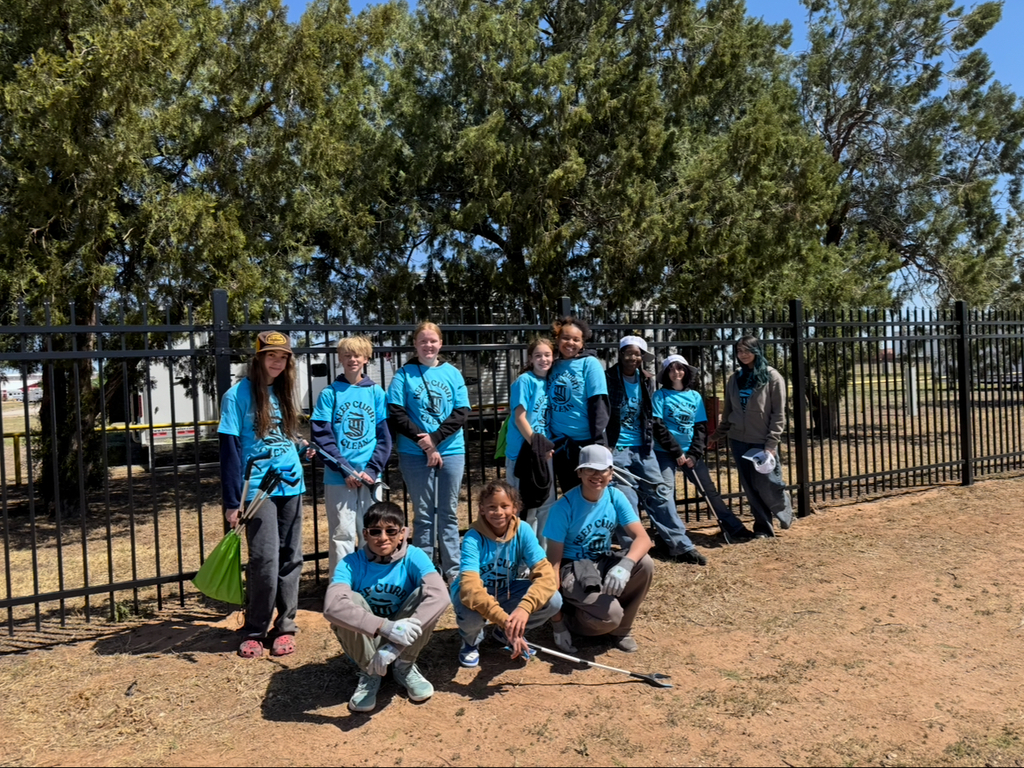 A group of people in blue shirts stands in front of a fence. Some hold gloves and tools. Trees and dirt are in the background.