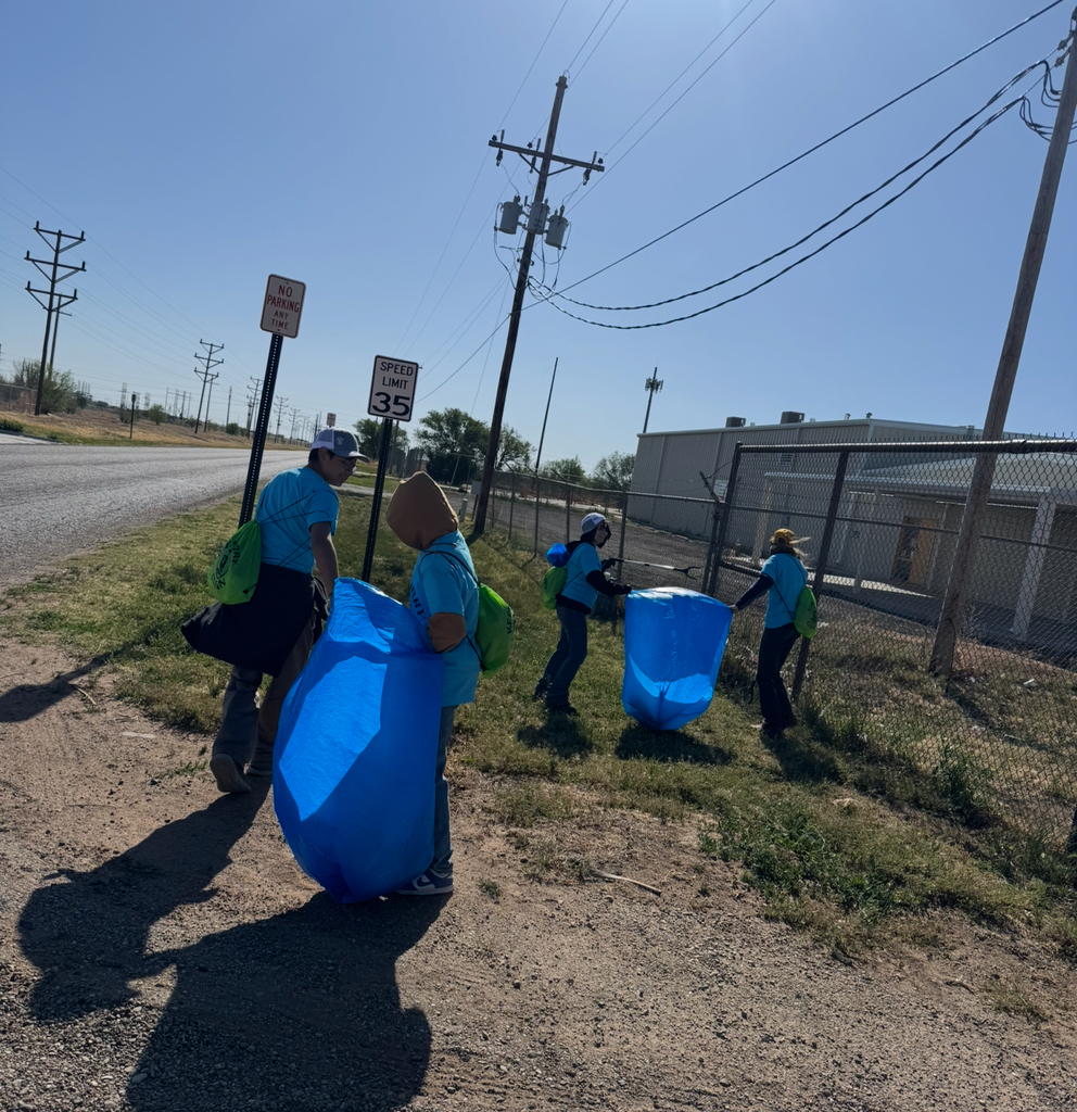 Four people are carrying blue bags, possibly picking up litter on a dirt road. A speed limit sign is posted.