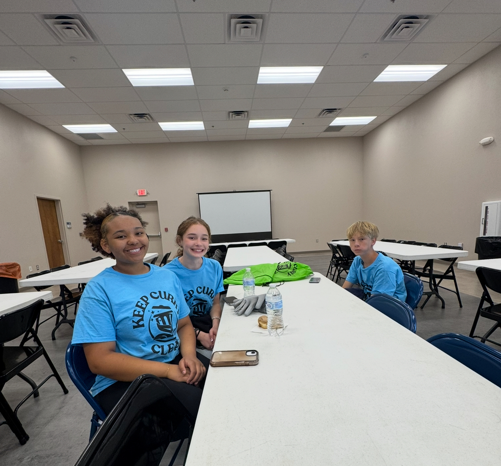 Three people in matching blue shirts sit at a table. One has a cell phone, and another holds a bag.