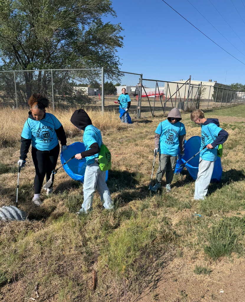 Group of people in matching shirts holding sticks and bags. One person has a blue bag. Background includes grass, tree, fence.