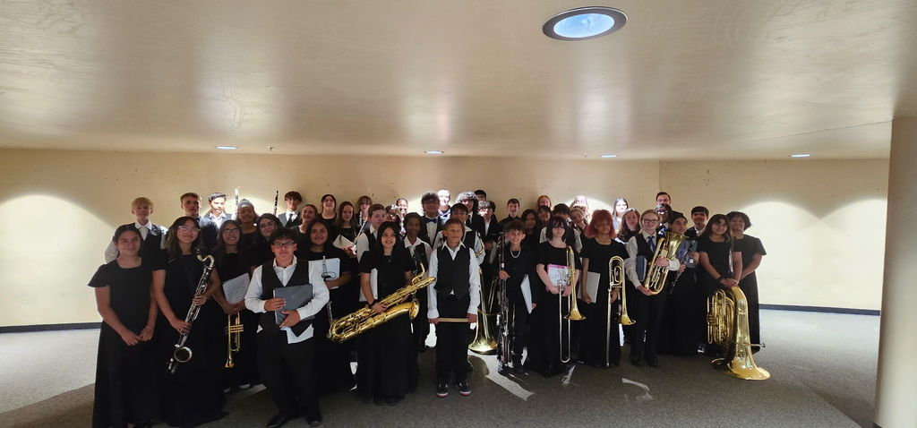A group of people in matching black attire, holding musical instruments, pose in a large room with white walls and ceiling.