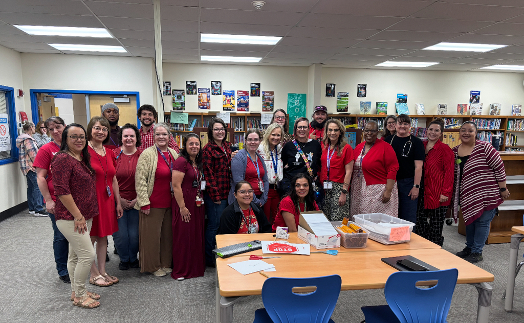 A group of adults in red shirts and dresses in a library. A table has papers and boxes on it.