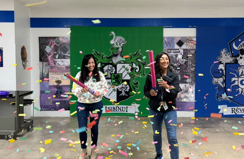 Two women with party blowers pose in a room with colorful confetti. A green banner with a crest is behind them.