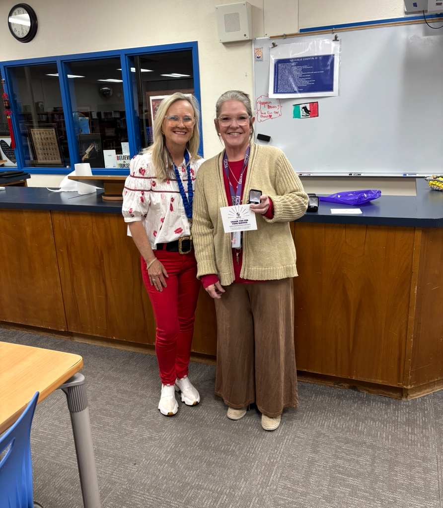 Two adults standing behind a counter in a classroom. One holds a paper with a logo.
