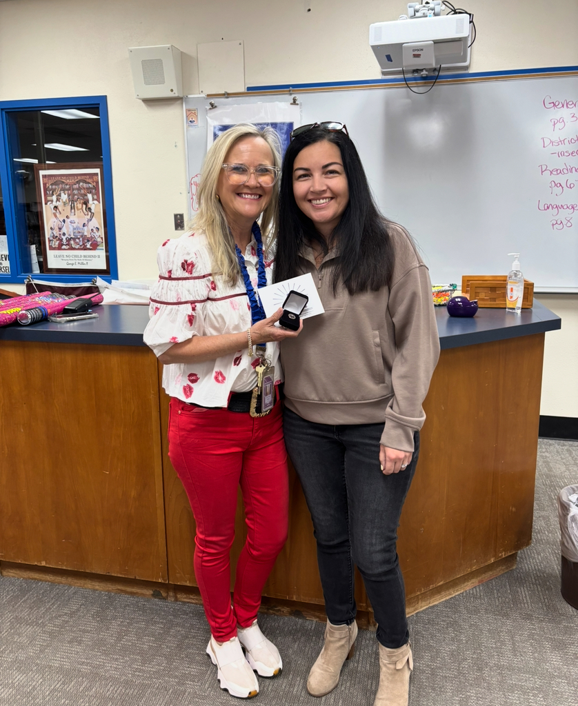 Two women stand in front of a counter, one holding a medal, both smiling. Behind them, a whiteboard is visible.