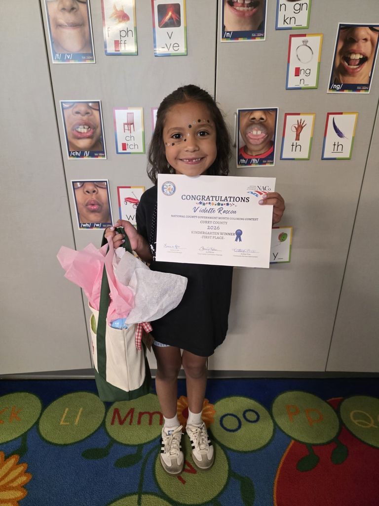 A young child holding a certificate for 'Student of the Month' in front of a wall with alphabets.