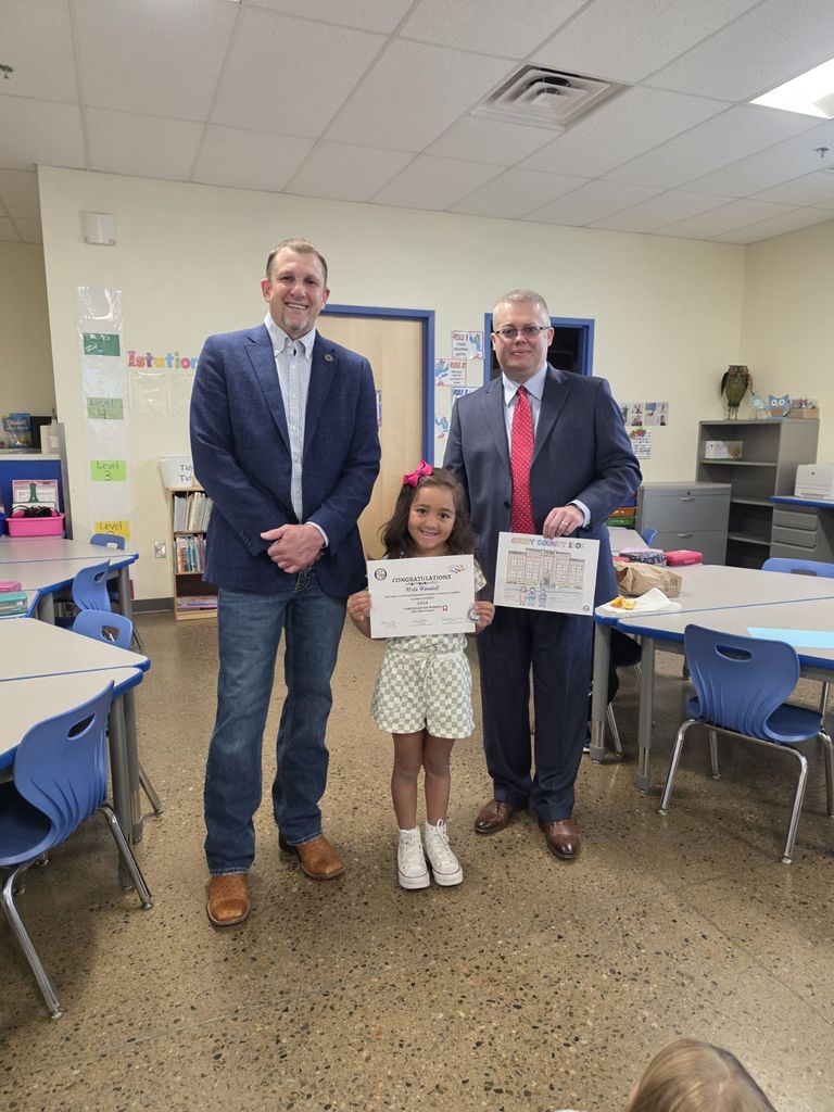 Two men and a young girl hold certificates in a classroom with desks, chairs, and bookshelves.