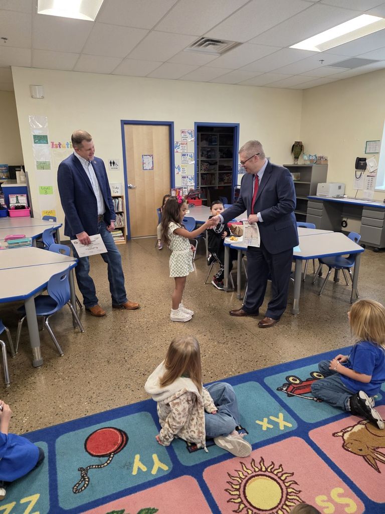 Two men in suits stand in a classroom, one giving a paper to a young girl. Children sit on a rug with letters and pictures.