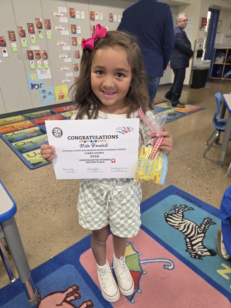 A girl holds a certificate and a gift bag. She stands in a classroom with colorful carpets and bulletin boards.