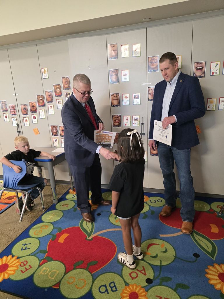 Two men and a young girl stand inside a room. One man hands a certificate to the girl.
