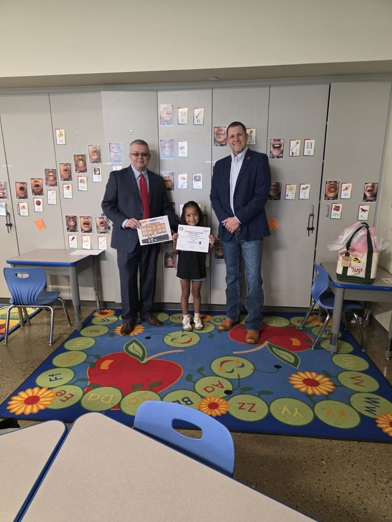 Two men and a young girl hold a certificate inside a classroom. The girl stands between the men.