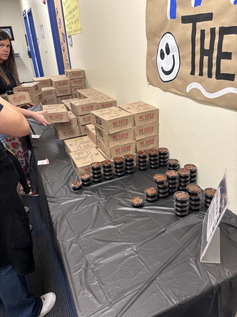 A table covered with a black tablecloth displays food in small containers. Behind the table, a banner says "The Happy School" with a smiley face.