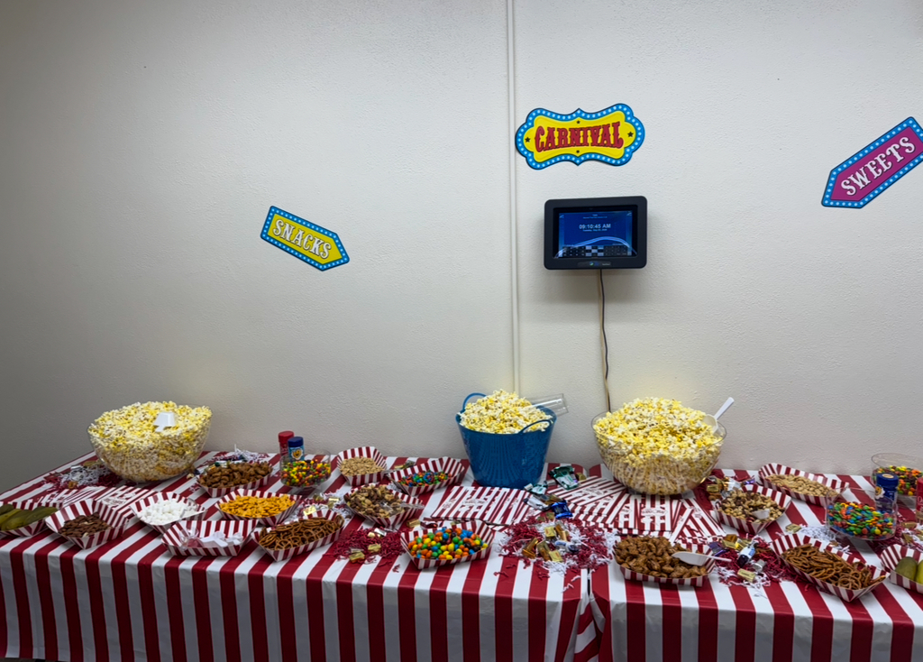 A table with a red and white striped tablecloth, featuring various candies and popcorn in bowls. Behind it, a wall has signs for 'Carnival' and 'Sweets'.