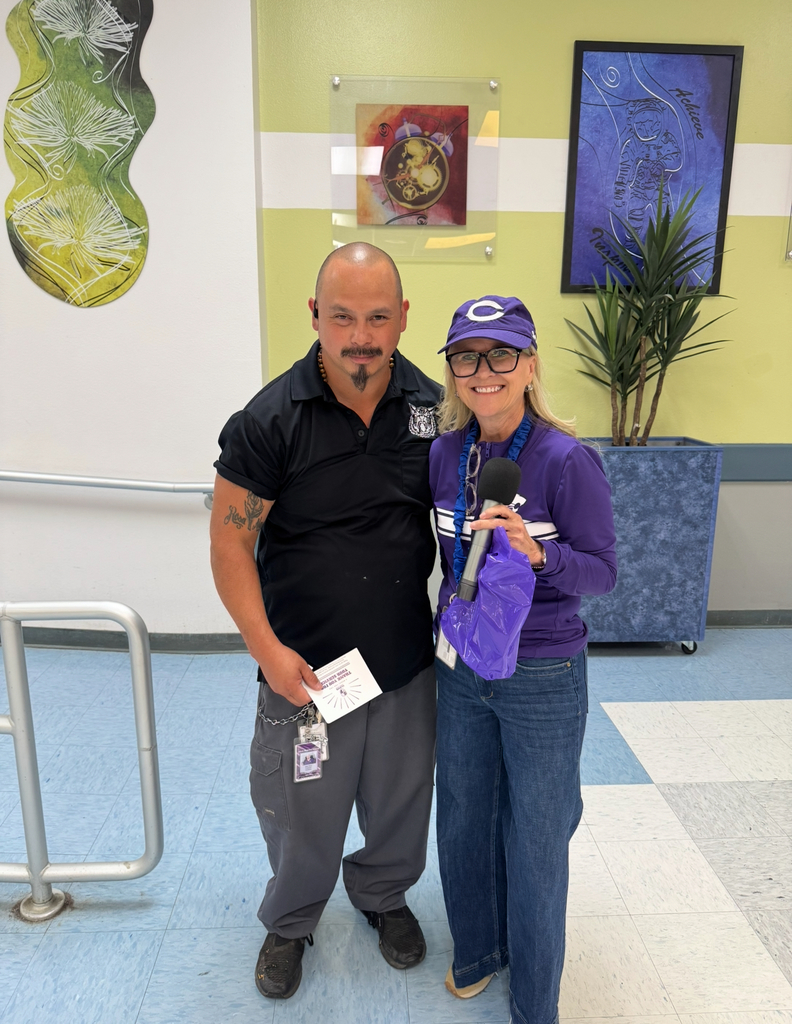 A man and woman stand in a hallway. The man wears a black shirt. The woman wears a purple shirt and holds a microphone.