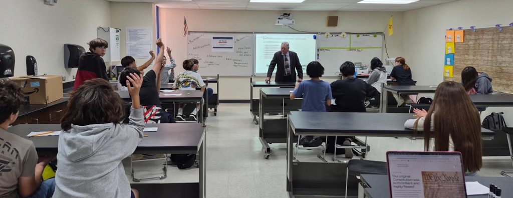 A classroom with students seated at desks. A teacher stands at a whiteboard, facing the students. The room has a bulletin board and various items on the walls.