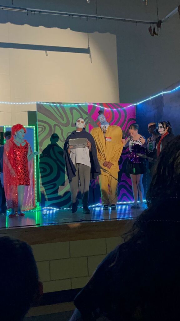 Actors on a stage, in various colorful costumes, with a backdrop featuring abstract patterns and lights.