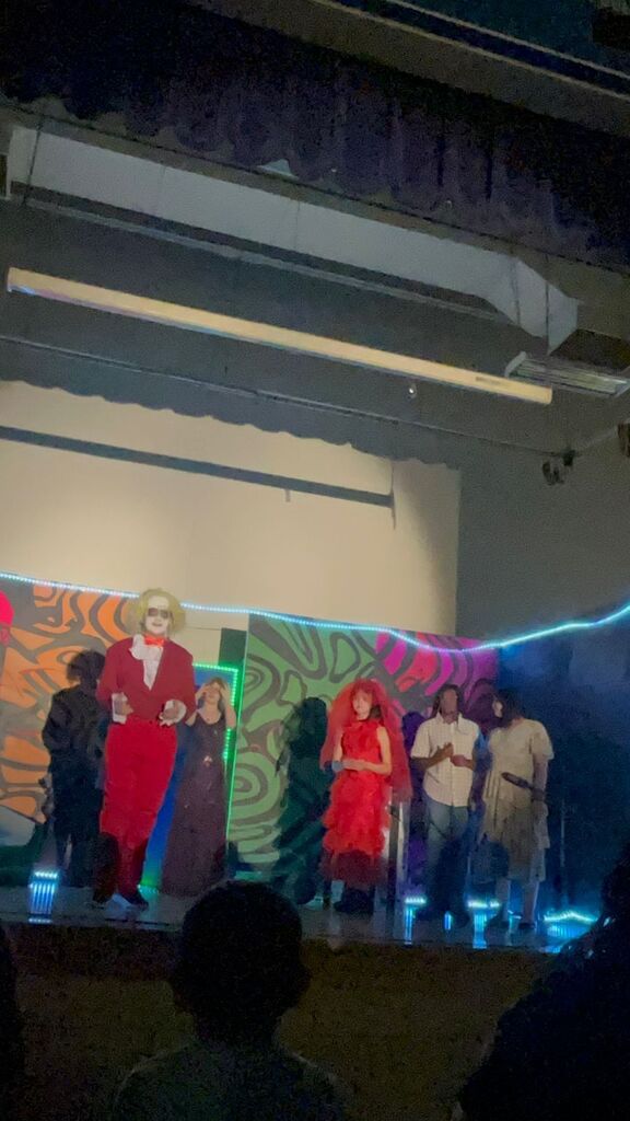Several people in various costumes on a stage with colorful backdrops. They are lit by blue and red lights.