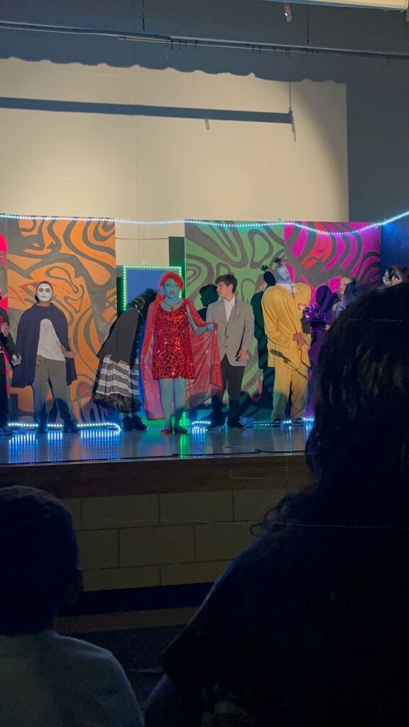 Performers on a stage in colorful costumes under a backdrop with abstract designs and lights.