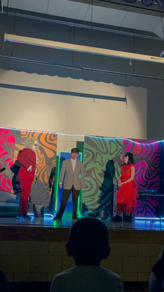 Three people on a stage wearing red and gray outfits with colorful backdrops. 