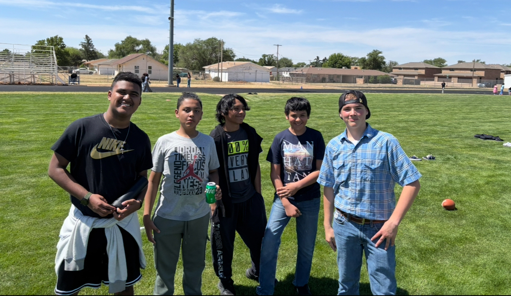 Five individuals stand in a grassy field with blue sky and distant trees. Each wears different clothing.